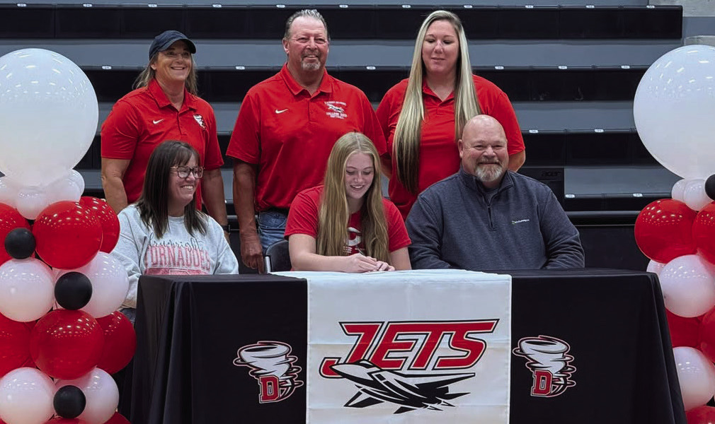 Evie Ools signs her letter of intent to play softball for the NOC Jets next to her mom, Megan Ools, and her dad Cary Ools. Behind her stood current coaches Alyssa Fuxa and Lesli Robinson, and future coach Steve Kessel.